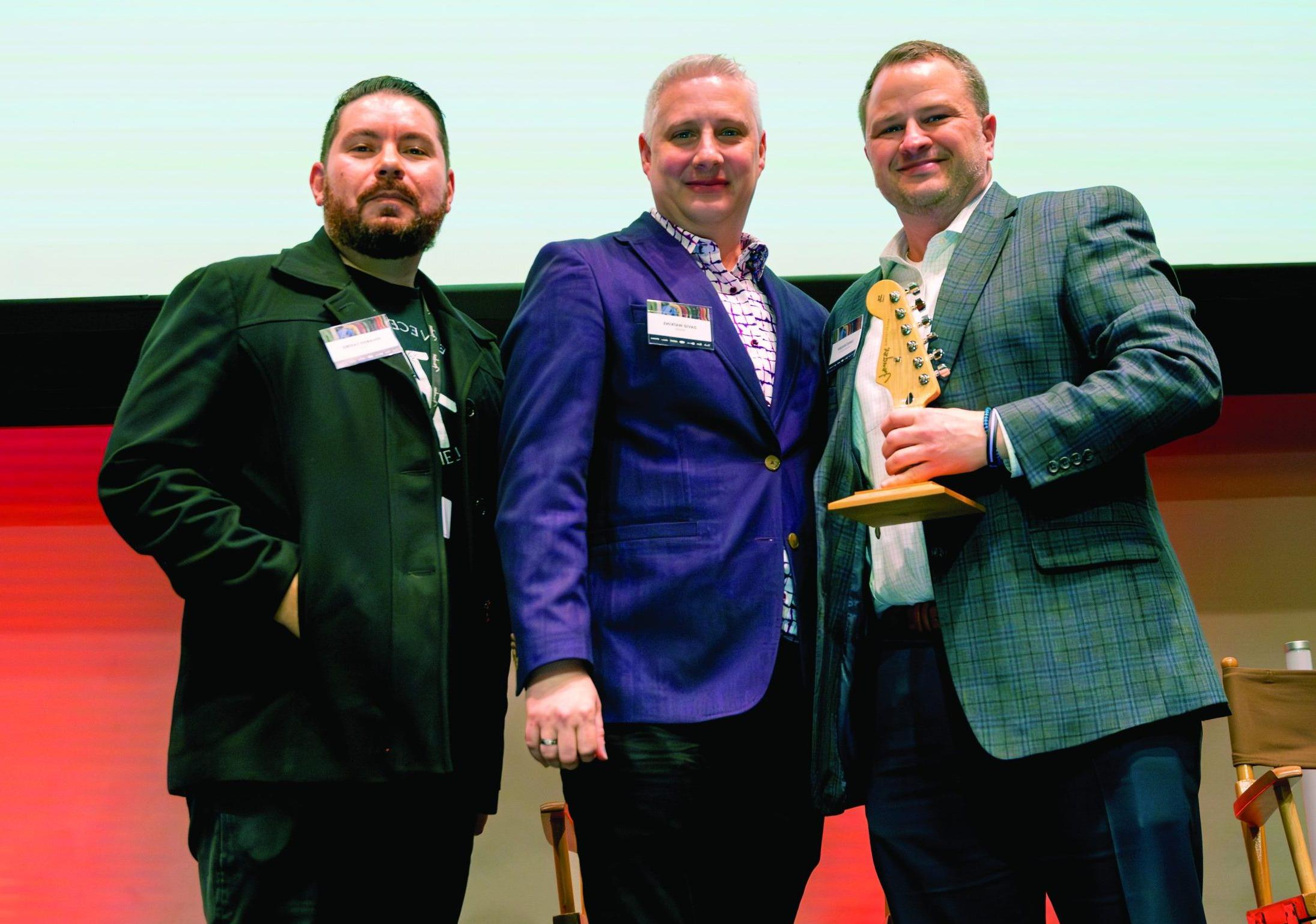 Three men post for a picture with the Pathfinder Award, the tuning pegs of an electric guitar.