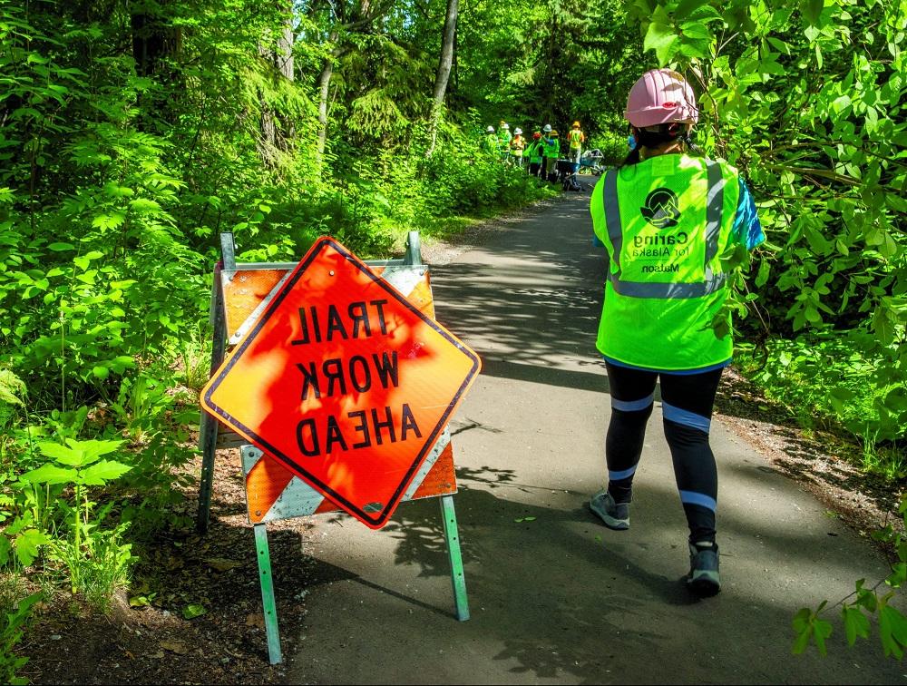 An Anchorage Park Foundation’s Youth Employment in Parks participant wearing a yellow Caring for Alaska vest walks past a Trail Work Ahead sign on a paved park path.