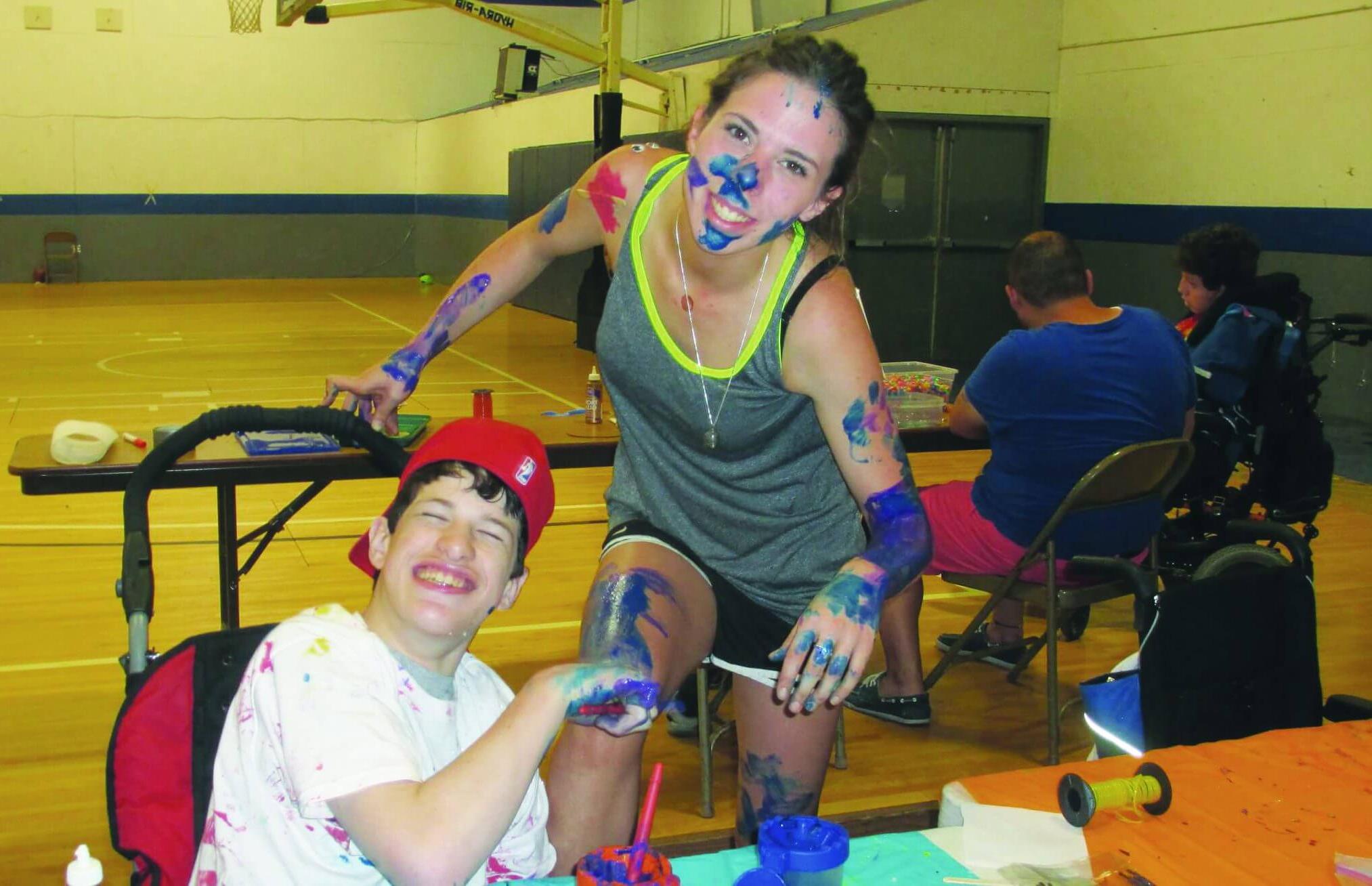 A happy camper wearing a red working with paint sits at a table while supervised by a counselor covered in paint from head to legs.