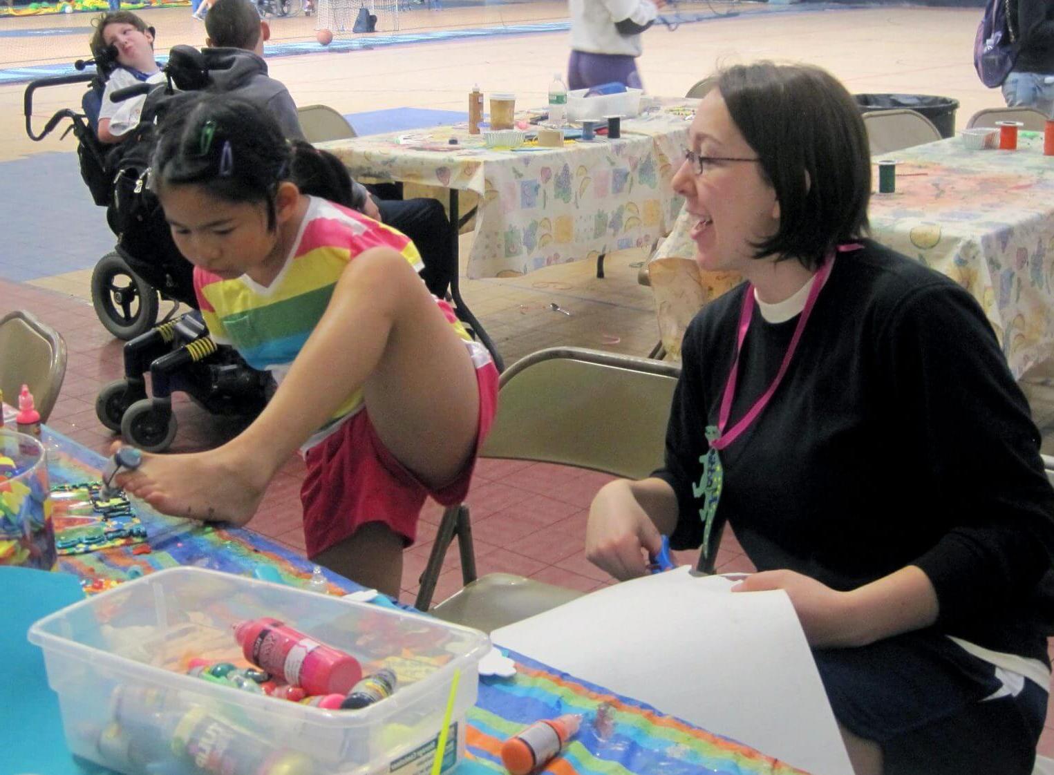 A young camper wearing a striped shirt uses her feet to complete a craft project while a counselor cuts paper.