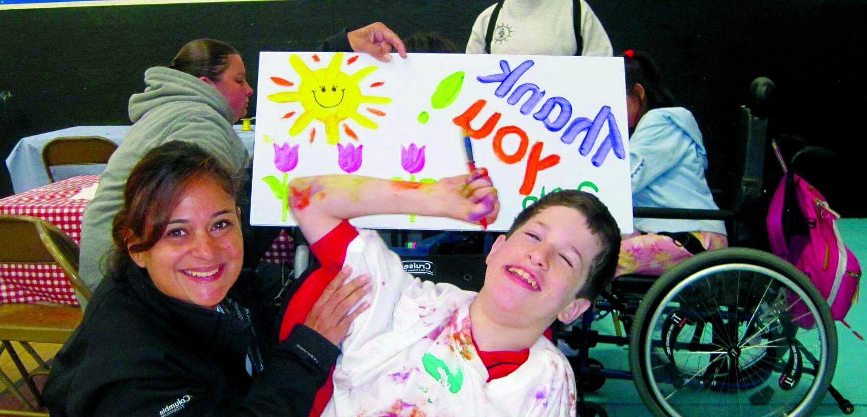 A happy camper wearing a white t-shirt splattered with pain holds a paint brush, while a counselor holds up a painted "thank you" poster with the sun and flowers.