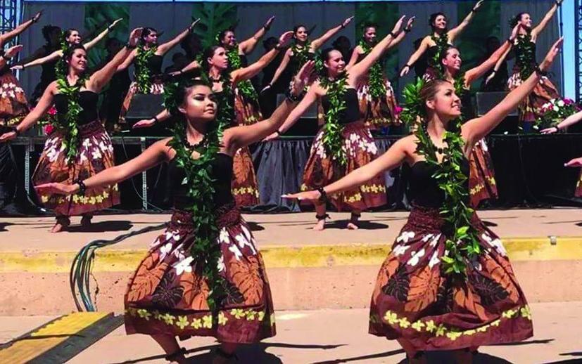 Female dancers adorned with ti leaf leis and brown costumes dance on stage.